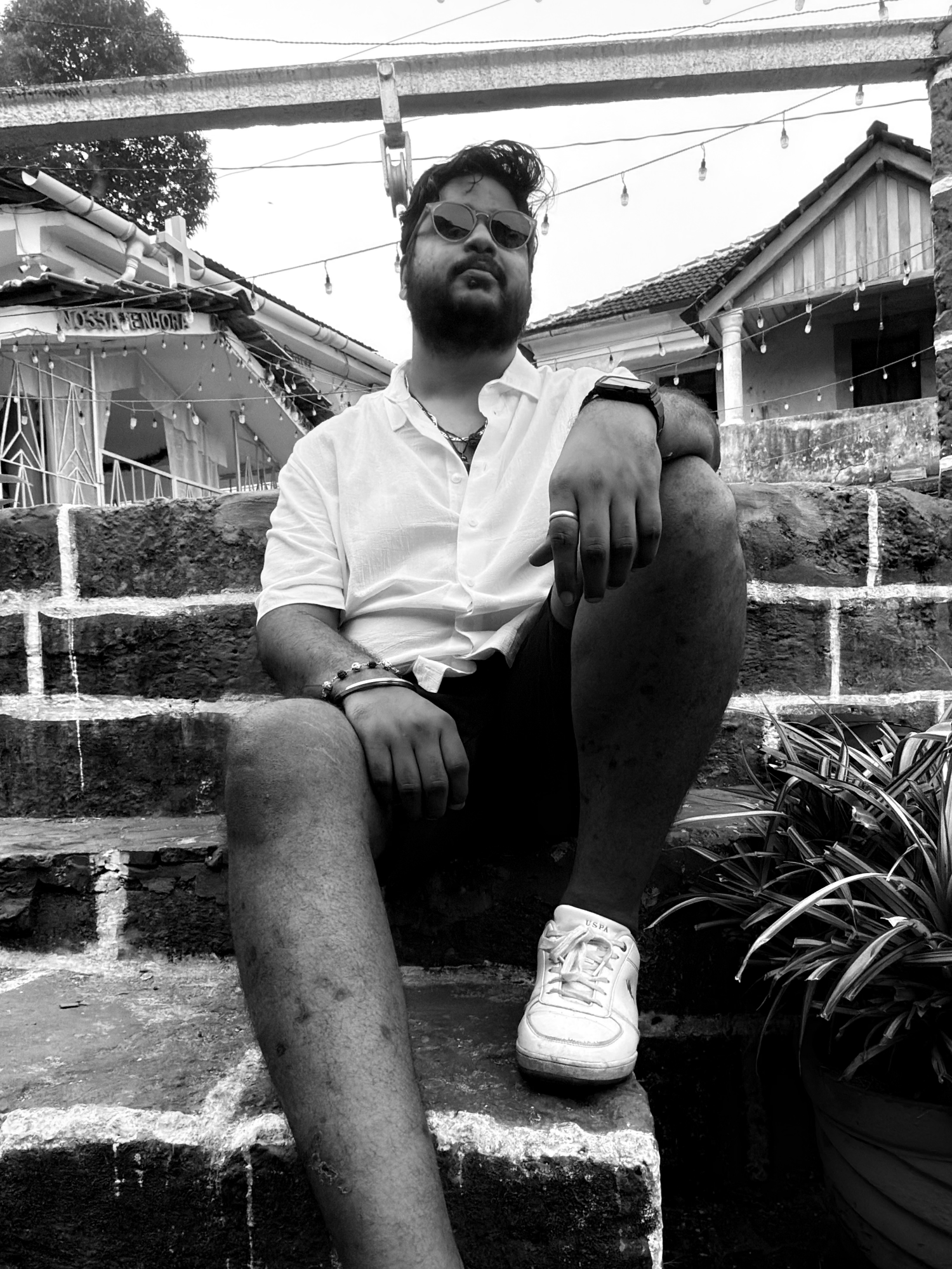 Abhishek Kumar sitting on stone steps in a white shirt and sunglasses, string lights and a tiled-roof building behind him. Black and white.